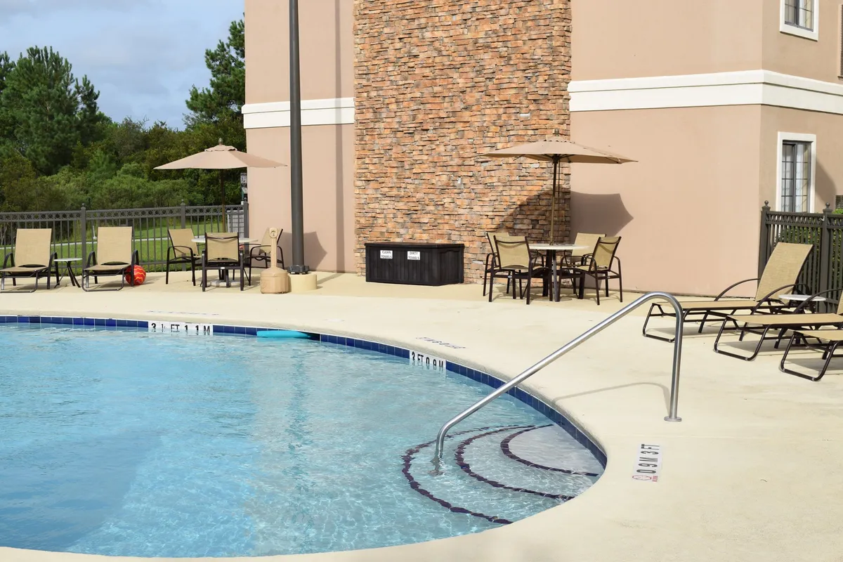 This exterior image features a sunlit swimming pool surrounded by lounge chairs and patio umbrellas, with a stone wall adding a touch of privacy. The inviting pool area is set against the backdrop of a pleasant outdoor space, where guests are seen swimming, sunbathing, and socializing. The mood is lively, capturing the sense of relaxation and recreation available on-site.