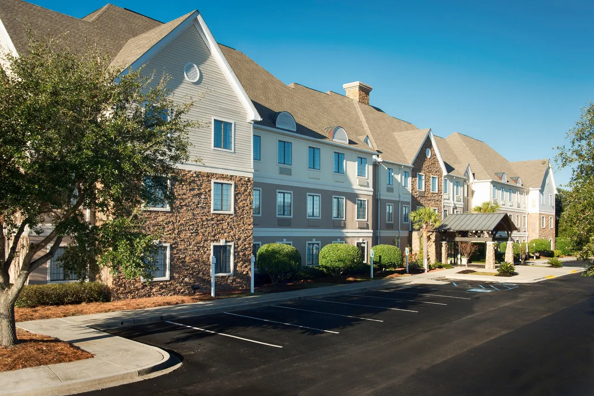 The image captures the exterior of the hotel with a welcoming entrance canopy, lush landscaping, and a clearly visible parking area for arriving guests. The scene suggests the first impression upon arrival and highlights the ease of access for travelers, including those with luggage or after a long journey.