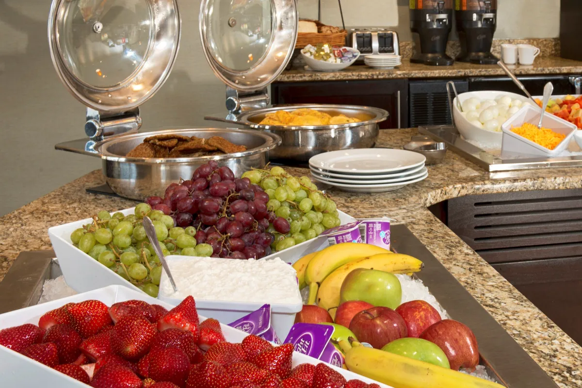 This image shows a close-up of a breakfast buffet with trays of hot food, a fresh fruit display, and coffee dispensers. The cheerful mood and focus on the food make it ideal for illustrating the quality and variety of the complimentary breakfast offerings.