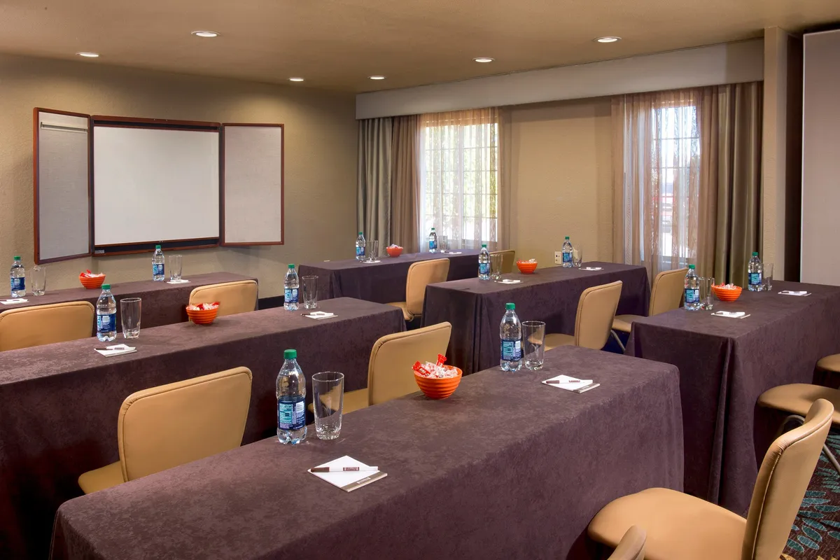 This image features a relaxed meeting room setup, with meeting tables arranged for group discussions, water bottles and snack bowls on the tables, and a presentation board in the background. The mood is focused but comfortable, implying a space suitable for business meetings, remote work, or collaborative projects.