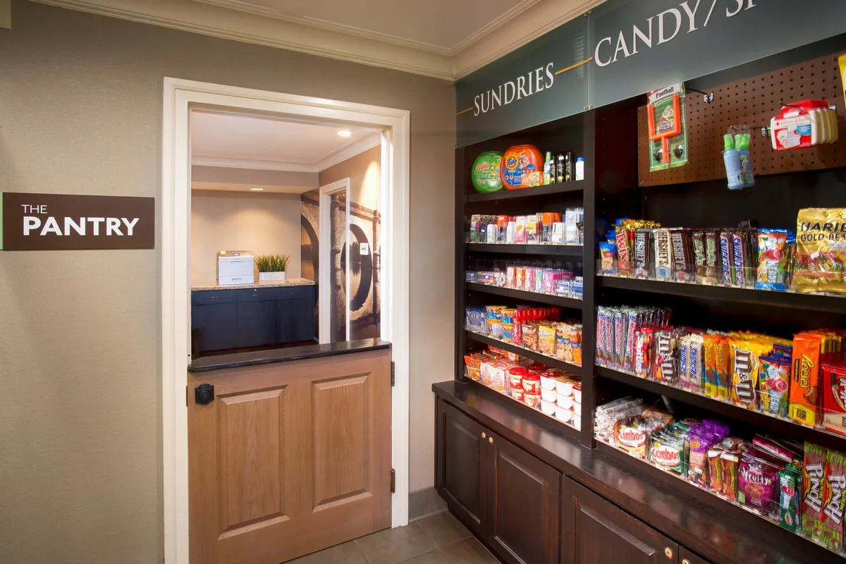 This image depicts 'THE PANTRY' area inside Staybridge Suites, with shelves filled with a variety of snacks and drinks. There is a visible entrance door, and the scene is warmly lit and welcoming, inviting guests to peruse and grab items as needed, suggesting a sense of convenience and accessibility for extended stays.