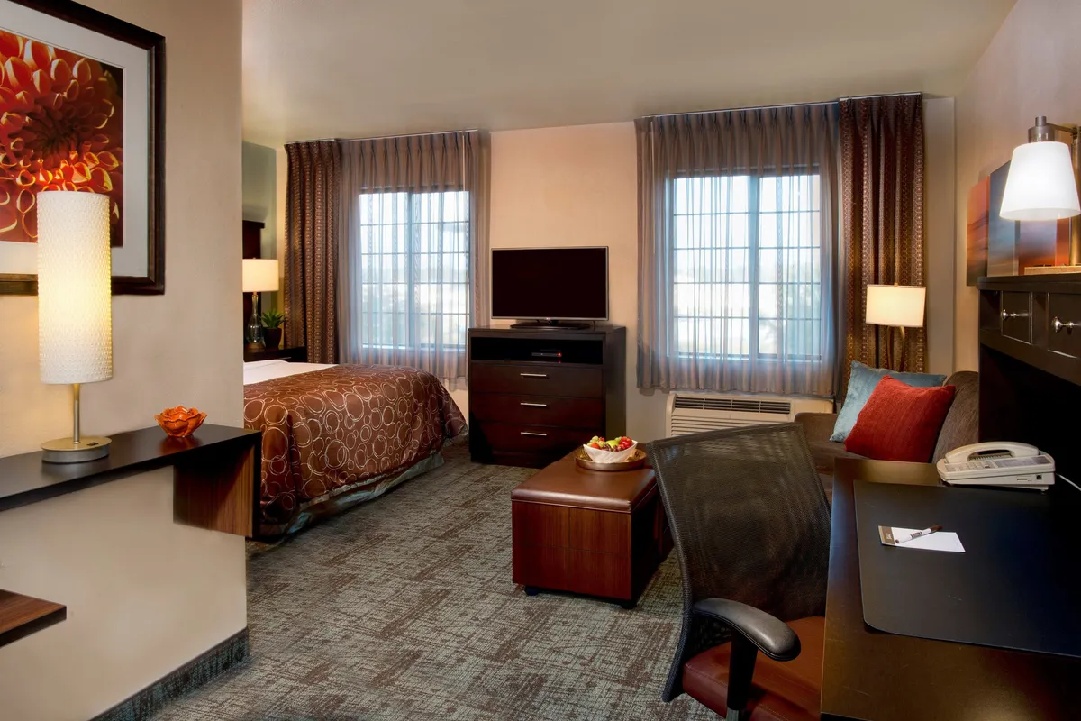 The image showcases a sunlit studio with a comfortable queen bed adorned with decorative bedding, a sleek desk and office chair perfect for getting work done, a flatscreen TV on a modern cabinet, a window dressed with soft curtains letting in natural light, and a table topped with a vibrant fruit bowl. The overall scene feels inviting, blending spaces for rest, work, and relaxation in a cohesive, relaxing atmosphere.
