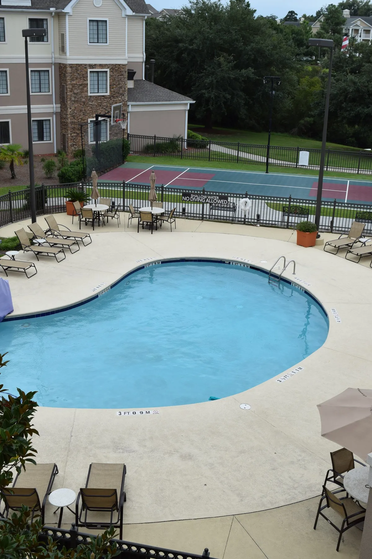 An inviting exterior image showcasing a sparkling swimming pool surrounded by lounge chairs and a nearby tennis court. The scene evokes relaxation and activity, with spaces for both sunbathing and playing sports, all set in a leisurely, sunlit environment.