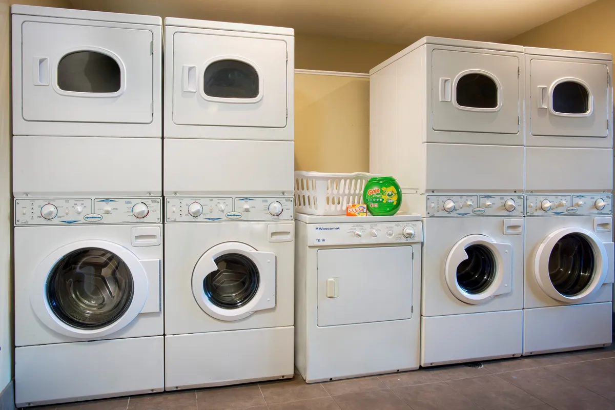 A tidy laundry facility with stacked washer and dryer units, a laundry basket, and detergent supplies is shown. The area is brightly lit, organized, and designed for guest convenience, making it easy to keep clothes fresh during both short and extended stays.