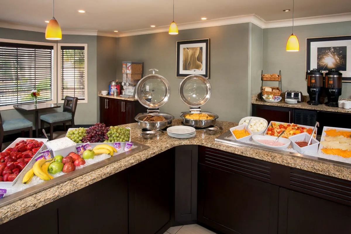 This image features a buffet table set with a colorful fruit display, coffee machines, and a welcoming dining area. The scene captures guests socializing and enjoying breakfast in a bright, communal space. The arrangement suggests both self-service convenience and a sociable atmosphere, aligning with the section's emphasis on complimentary breakfast and flexible dining.