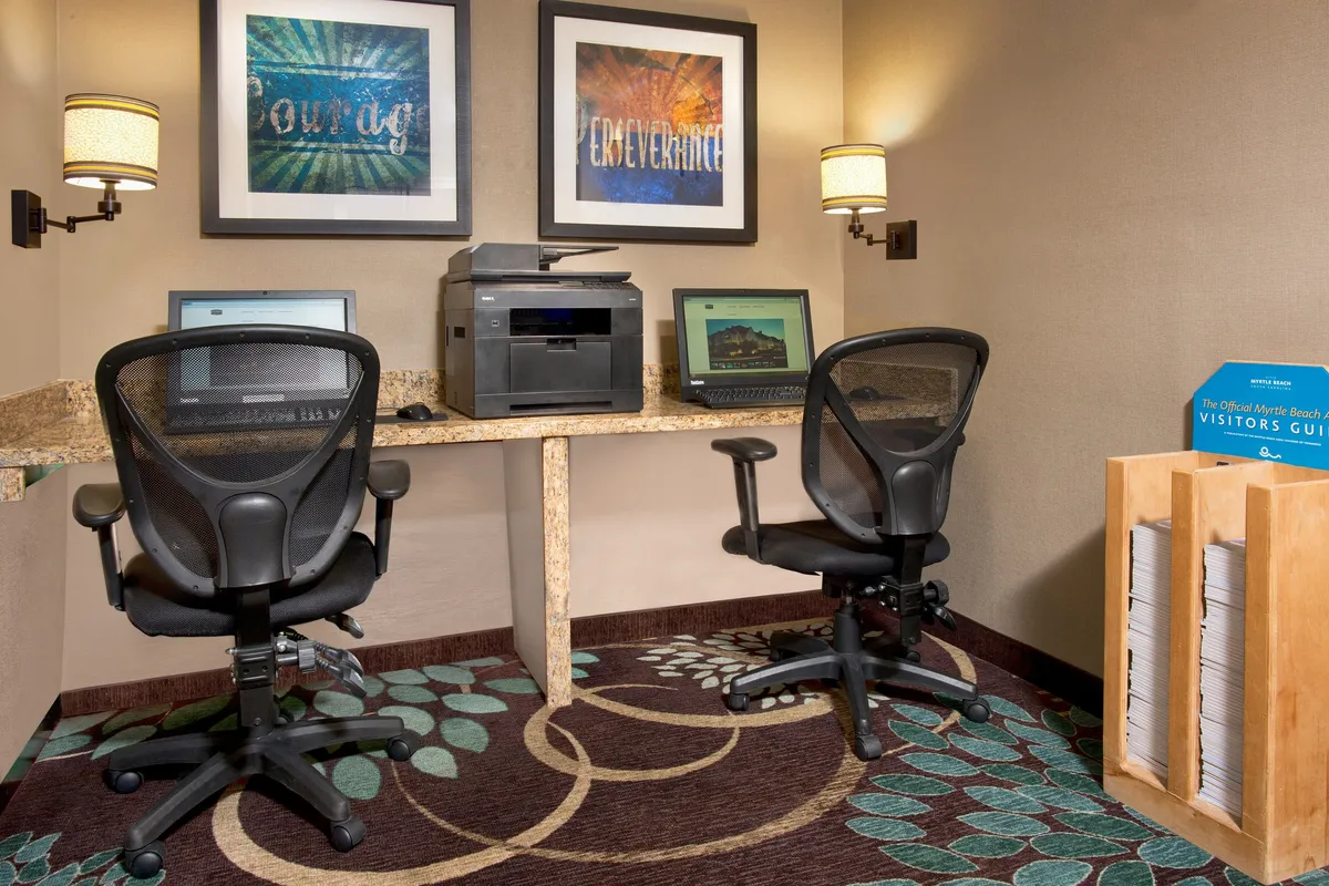 This interior image displays a well-equipped business center, featuring modern computers, a printer, motivational artwork, comfortable seating, and a magazine rack. The space is designed for productivity, with a welcoming environment that encourages guests to work, print documents, or plan activities.